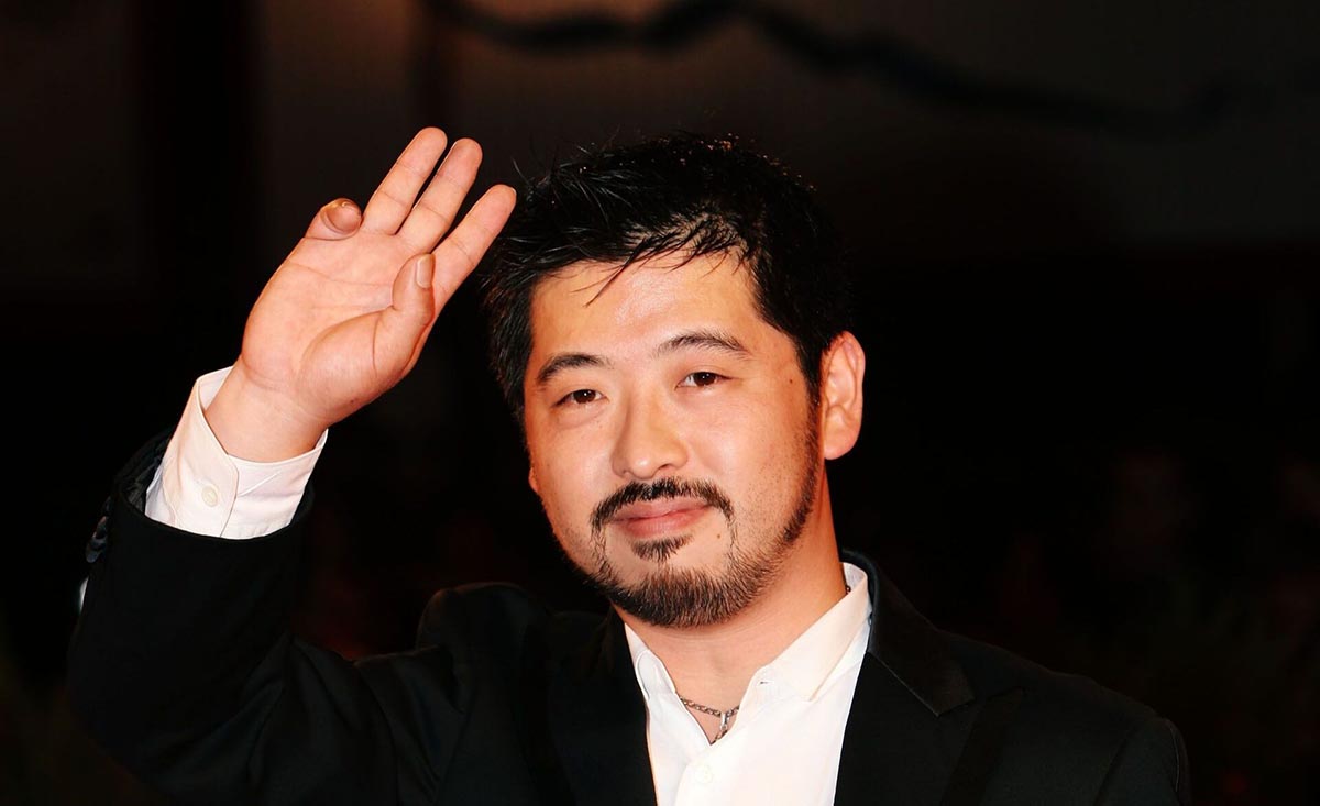 Takashi Shimizu dalam setelan jas hitam dan putih, sedang melambaikan tangan